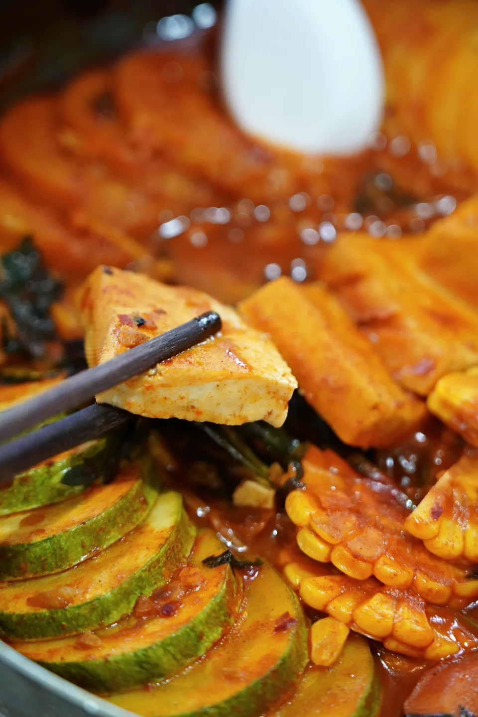 Chopsticks lifting tofu above vegetables in a pot of red broth