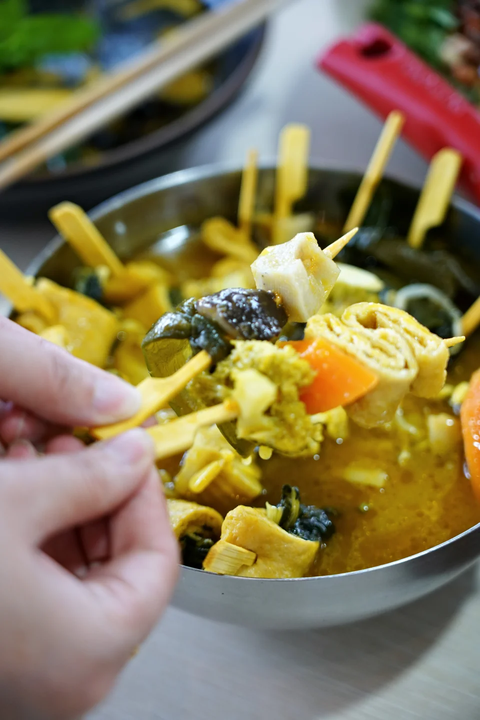 A hand lifting vegetable skewers from a pot of sauce